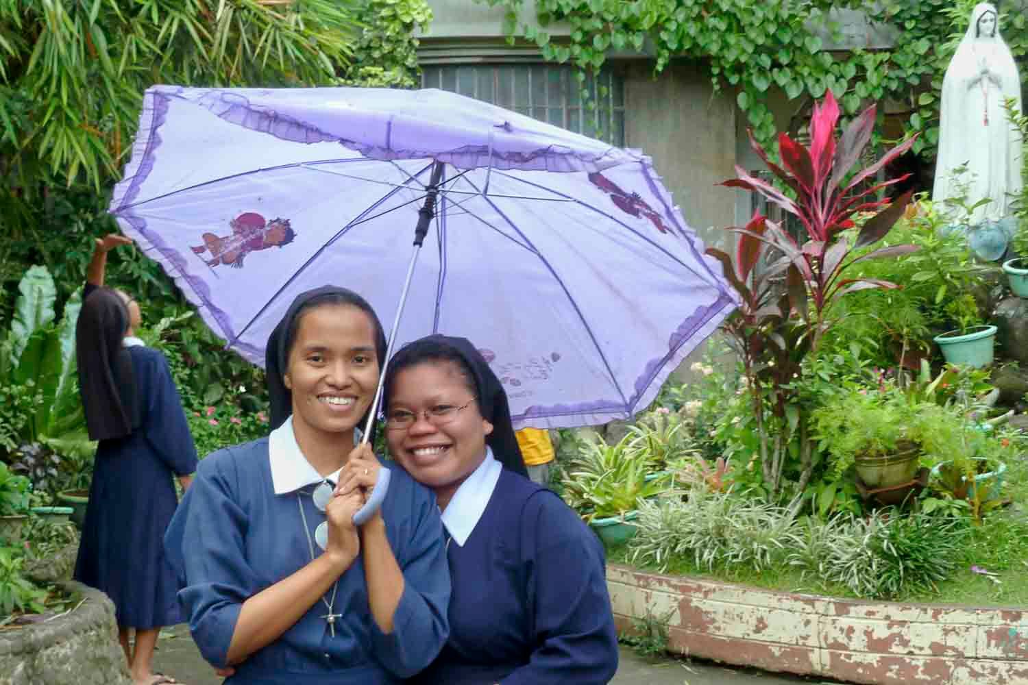 Zwei Nonnen lächeln und halten einen violetten Regenschirm in einem Garten mit einer Statue.