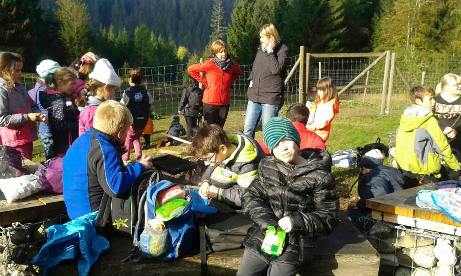 Kinder versammelten sich im Freien mit Erwachsenen; einige saßen, andere standen. Möglicherweise ein Schulausflug auf einer Wiese mit Bäumen.