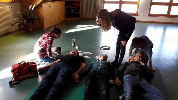 Menschen üben Erste Hilfe auf dem Boden; einer kniet neben einer Arzttasche, andere schauen zu.