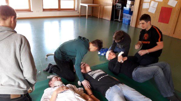 Eine Gruppe von Menschen übt Erste Hilfe an zwei Personen, die in einem Innenraum auf einer Matte liegen.