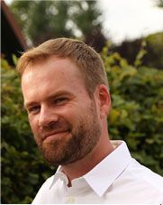 Ein Mann mit kurzem blonden Haar und Bart lächelt; er trägt ein weißes Hemd; im Hintergrund Laubwerk.