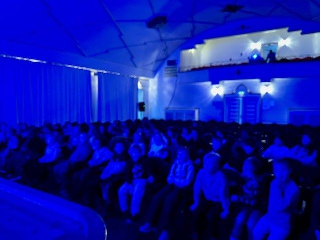 Publikum in blau beleuchtetem Theater; Bühne verdeckt. Die Zuschauer sitzen und blicken nach vorn.