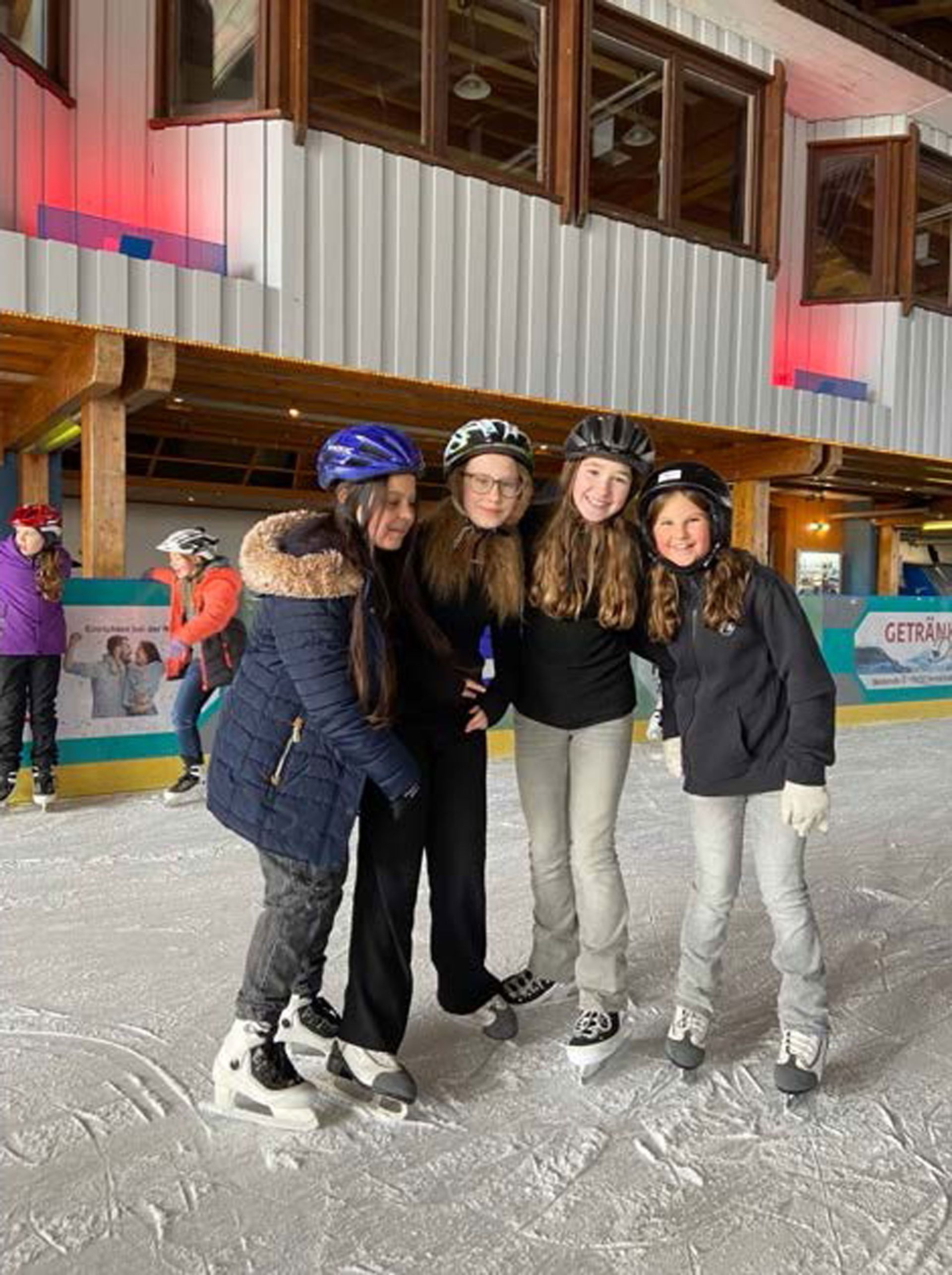 Vier Mädchen auf Schlittschuhen posieren lächelnd auf einer Eisbahn.