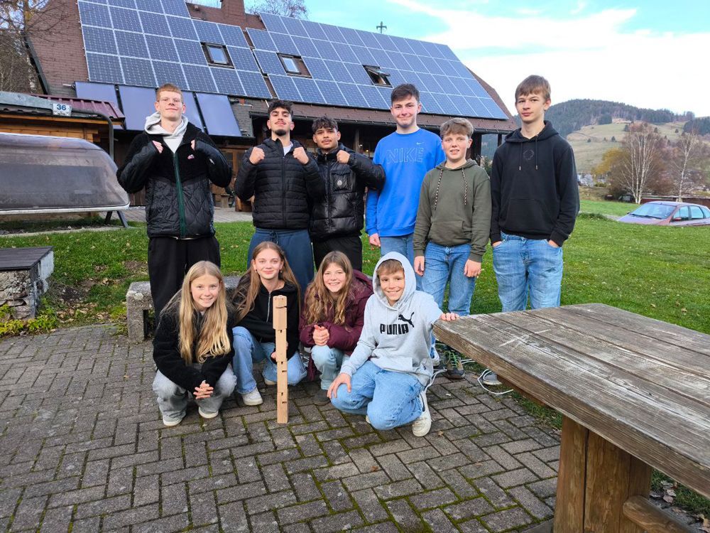 Eine Gruppe von Menschen steht im Freien in der Nähe eines Hauses mit Solarpaneelen. Einige lächeln und halten einen Holzstock.