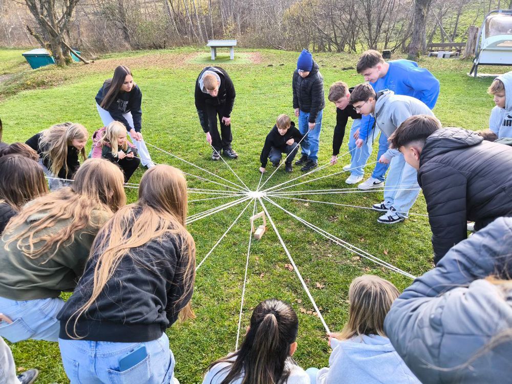 Eine Gruppe von Menschen spielt im Freien auf einer Wiese ein gemeinsames Spiel mit Schnüren und einem zentralen Gegenstand.