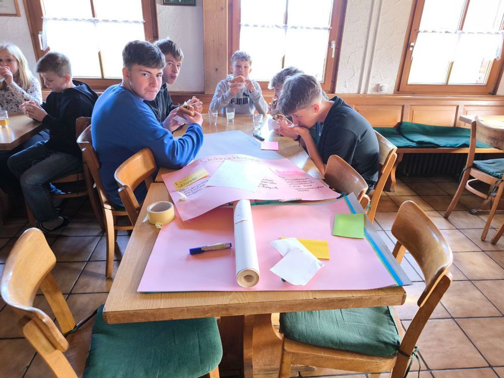 Eine Gruppe Kinder sitzt an einem Tisch und arbeitet mit farbigem Papier, Stiften und Haftnotizen an einer Landkarte.