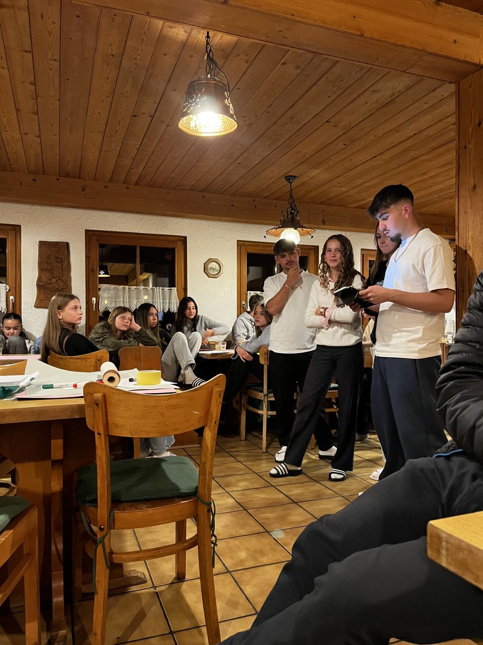 Die Leute hatten sich in einem rustikalen Speisesaal versammelt. Zwei Personen standen in der Nähe der Mitte und unterhielten sich, während andere an Tischen zuhörten.