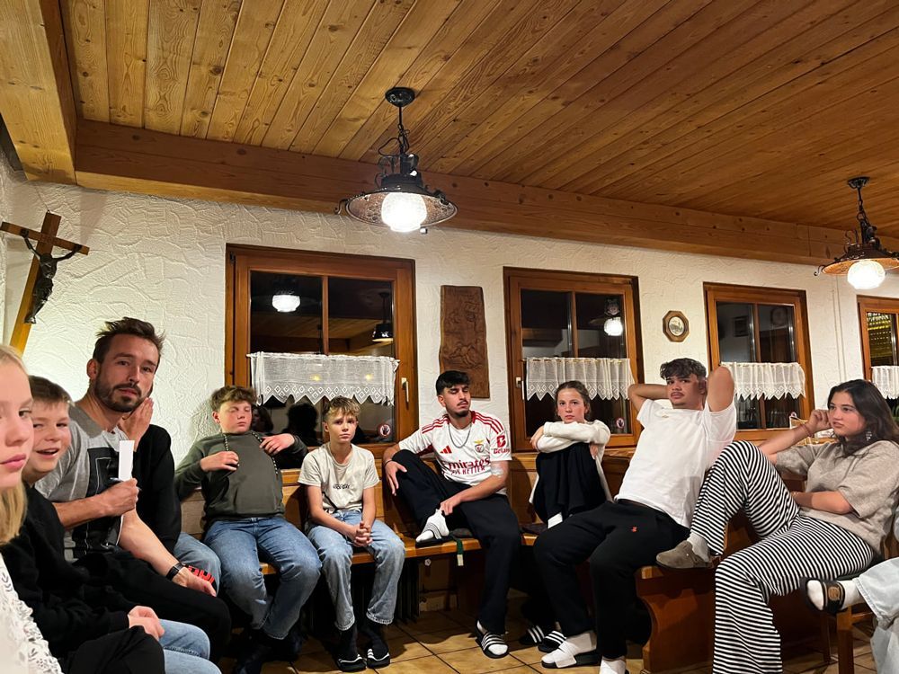 Menschen sitzen drinnen und hören zu; Holzdecke und -wände, Kruzifix, Fenster.