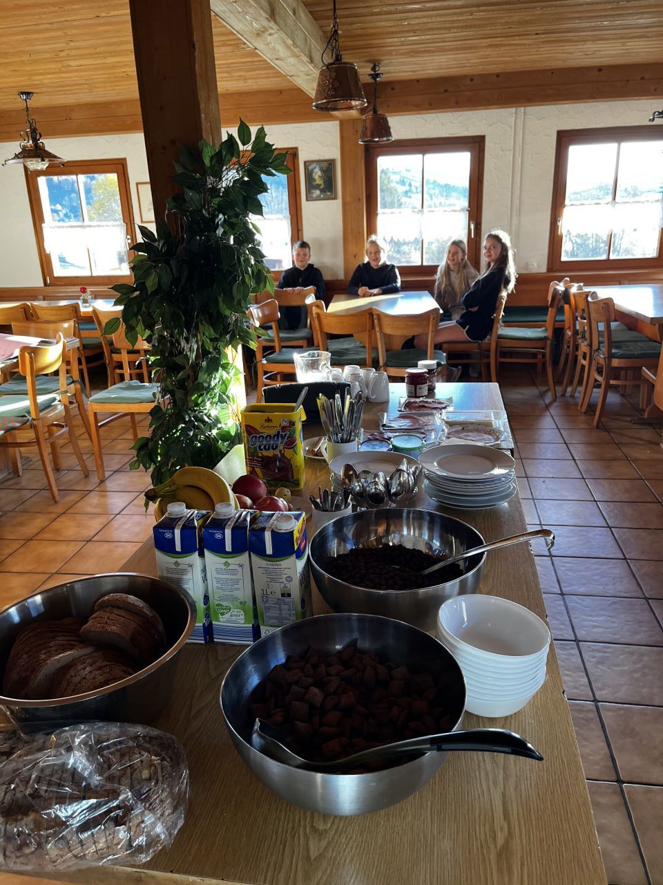 Frühstücksbuffet mit Brot, Müsli, Obst und Gästen, die an Tischen in einem rustikalen Speisesaal sitzen.