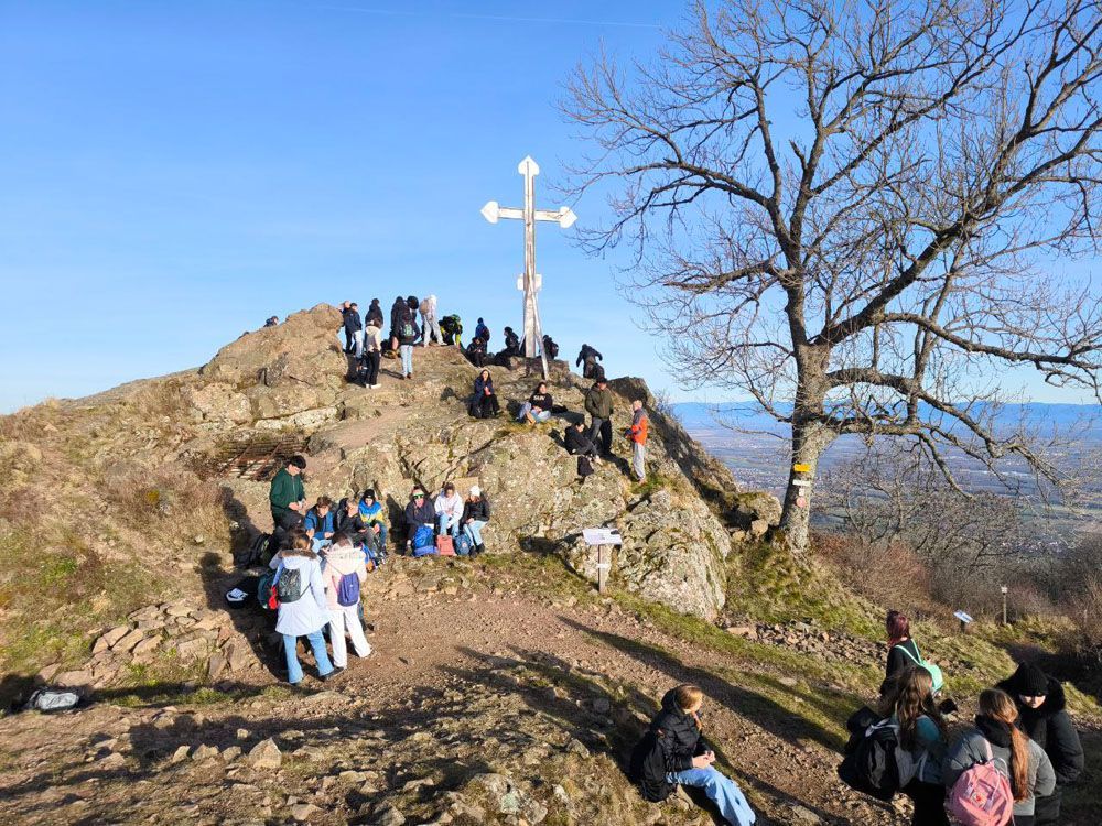 Menschen versammeln sich auf einem felsigen Hügel mit einem großen Kreuz. Blauer Himmel, kahler Baum und ein Tal im Hintergrund.
