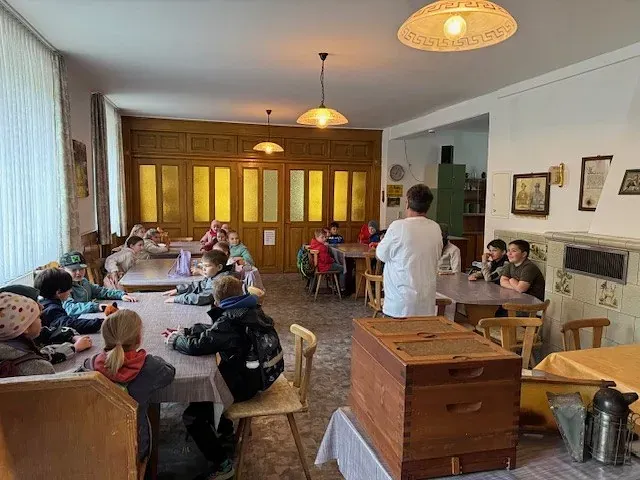 Kinder in einem Raum mit einer Lehrerin, Tischen und einem Bienenstock.