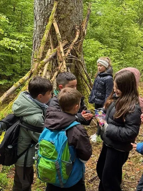 Kinder versammeln sich um einen Gegenstand im Wald. Eines hält etwas in der Hand. Stöcke lehnen an einem Baumstamm.