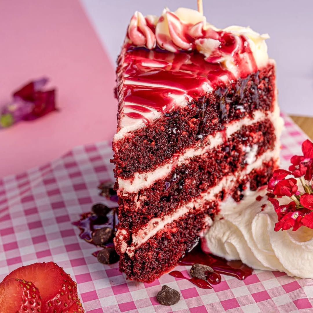 Rebanada de pastel red velvet con glaseado de crema y salsa de fresa sobre un mantel a cuadros rosa.