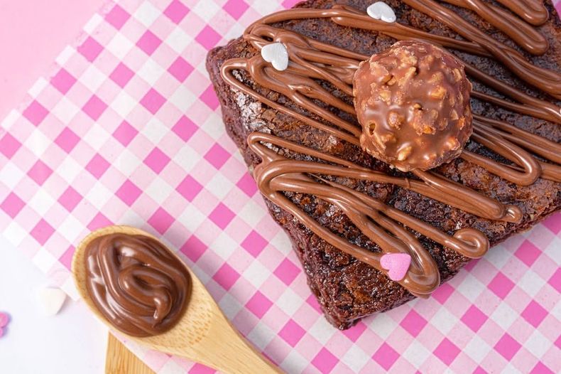 Brownie bañado en chocolate sobre papel rosa a cuadros con un bombón Ferrero Rocher y una cucharada de crema de chocolate.
