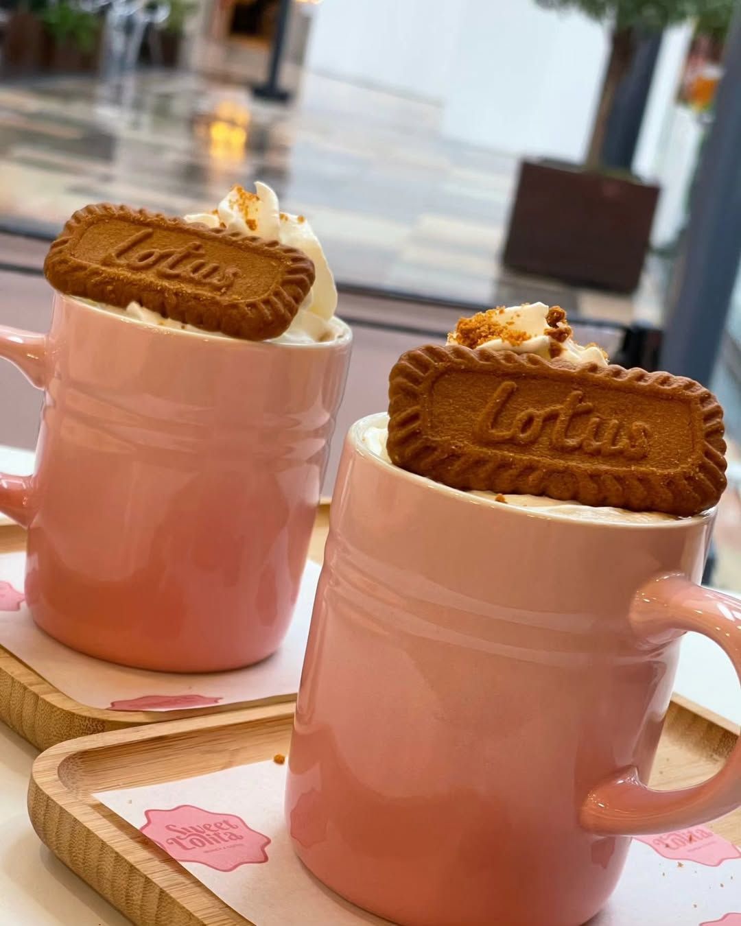 Dos tazas rosas con galletas Lotus encima, dispuestas en bandejas en una cafetería luminosa.