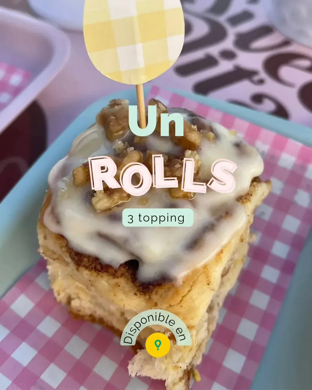 Rollo de canela con glaseado blanco sobre una superficie rosa a cuadros, con el texto 