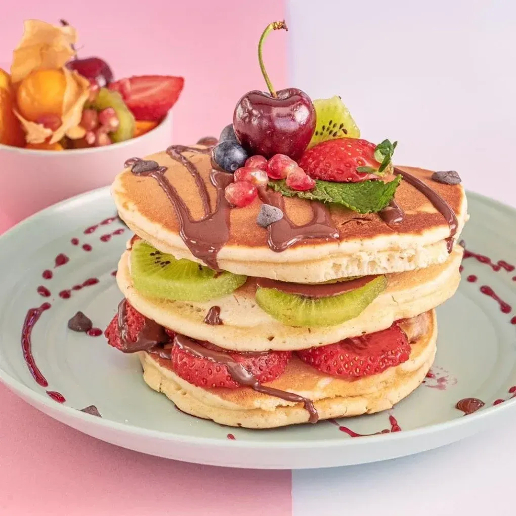 Pila de panqueques cubiertos con fruta, chocolate y crema batida en un plato de color pastel.