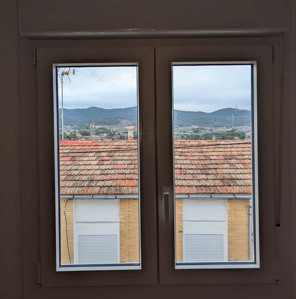 Vista a través de una ventana de tejados, edificios y montañas lejanas bajo un cielo nublado.
