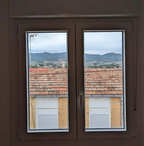 Vista a través de una ventana de tejados, edificios y montañas lejanas bajo un cielo nublado.