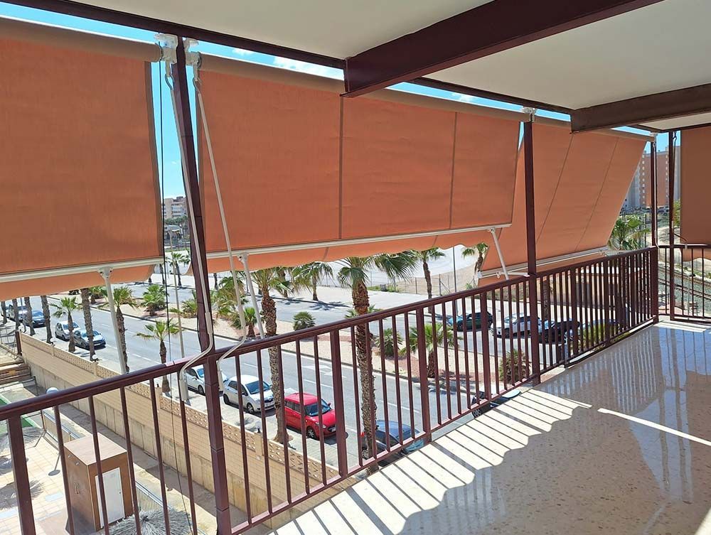Vista desde el balcón con persianas marrones. Calle y vehículos visibles. Día soleado.