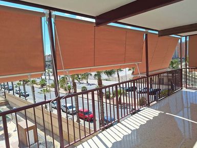 Vista desde el balcón con persianas marrones. Calle y vehículos visibles. Día soleado.
