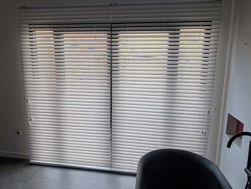 Ventana con persianas horizontales blancas cerradas. Silla oscura en primer plano.