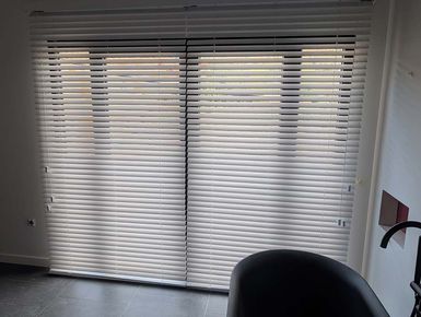 Ventana con persianas horizontales blancas cerradas. Silla oscura en primer plano.