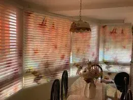 Habitación con persianas con motivos florales, lámpara colgante, mesa con flores y sillas.