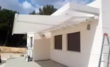 Un toldo blanco se extendía sobre las ventanas de un edificio, ofreciendo sombra. El edificio es blanco con marcos de ventanas marrones; se ve una escalera.