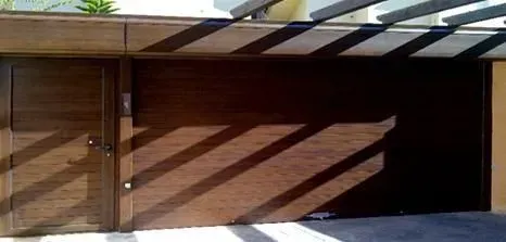 Puerta de madera y puerta de garaje bajo un techo de listones, proyectando largas sombras.