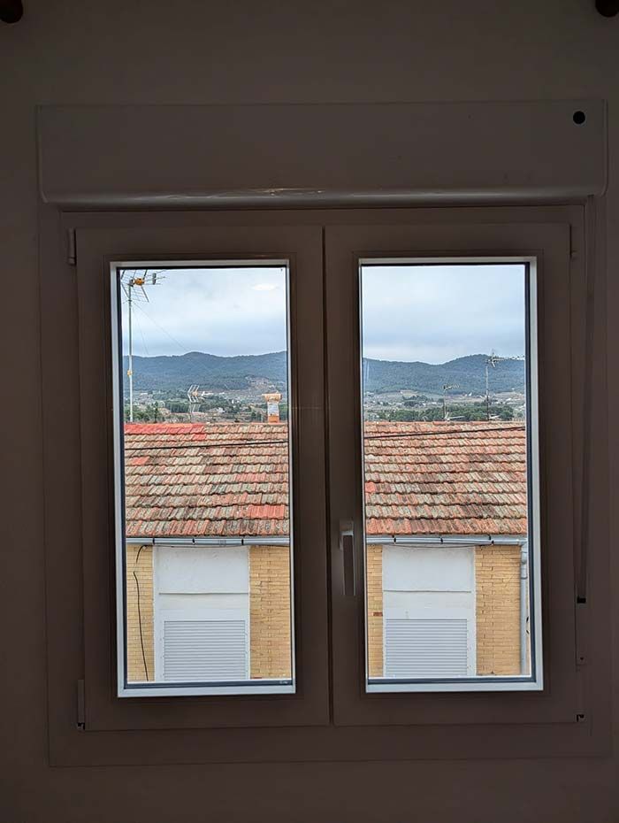 Ventana de dos paneles con vista a los tejados, a una ciudad lejana y a un cielo nublado.