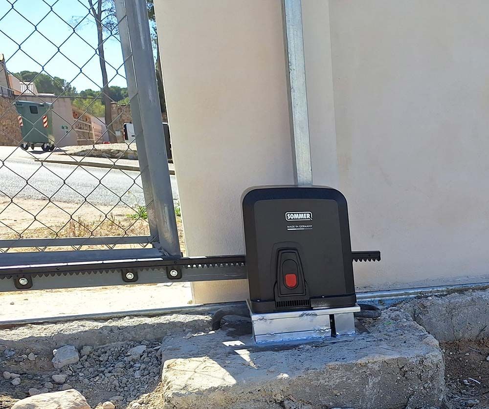 Motor para puerta corredera automática montado sobre base de hormigón, conectado a una valla de eslabones de cadena.