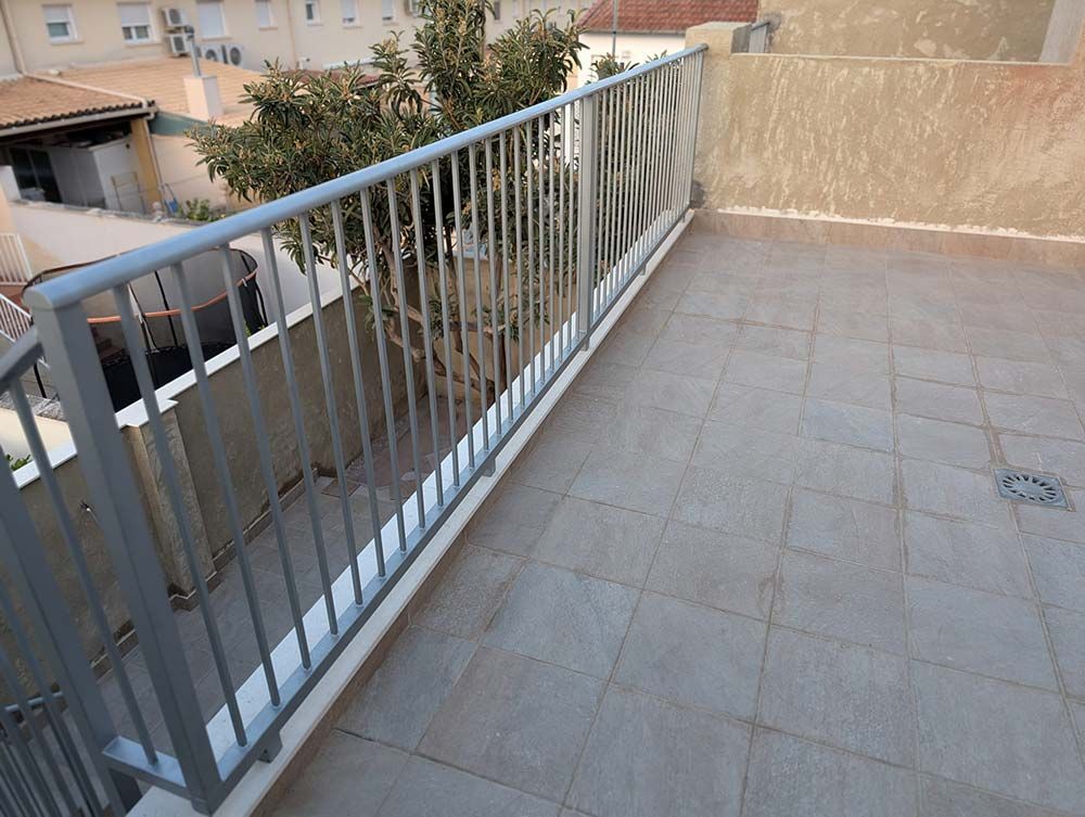 Barandilla de metal gris en un patio con azotea de tejas con un árbol visible al fondo.