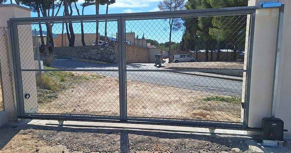Puerta corrediza metálica con malla de alambre, parcialmente abierta, dejando ver una carretera y edificios al fondo.