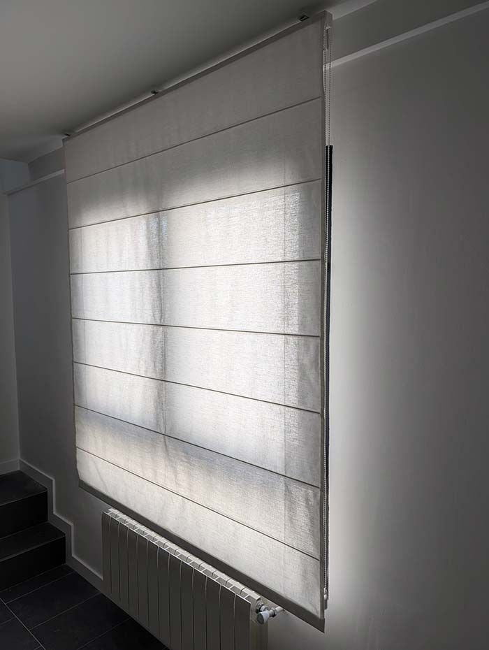 Una persiana romana blanca cubre una ventana y deja entrar la luz del sol. Junto a una pared con un radiador y un tramo de escaleras.