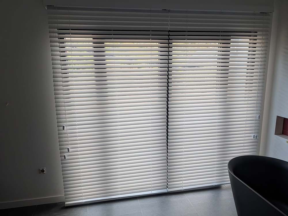 Persianas blancas cubren unas puertas de cristal, proyectando sombras horizontales. Se ve parcialmente una silla oscura.