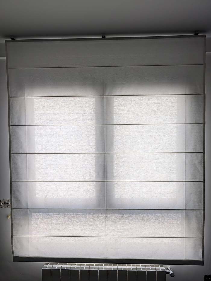 Una persiana romana cubre una ventana, mostrando pliegues horizontales, en una habitación con un radiador debajo.