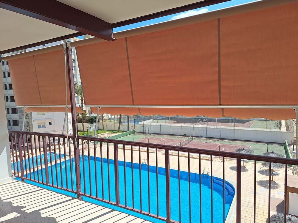 Balcón con vista a piscina y canchas de tenis, con cortinas marrones parcialmente rebajadas.