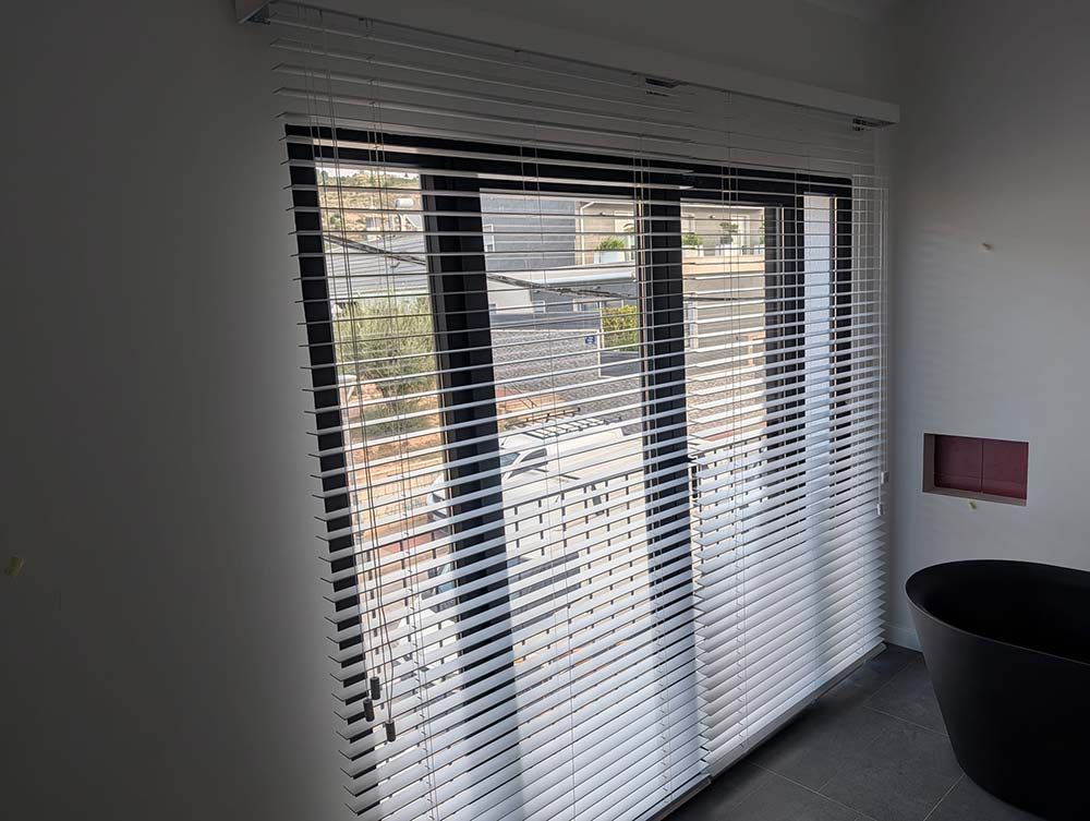 Ventana con persianas blancas, con vistas a los edificios. Hay una bañera negra en la esquina.