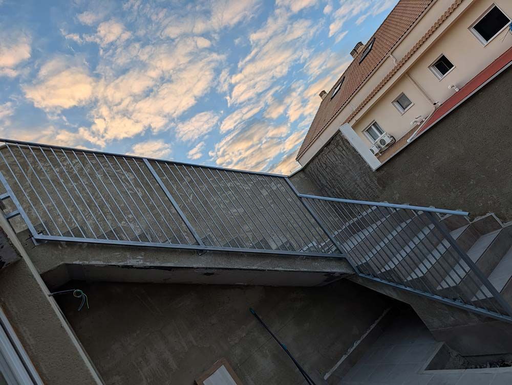 Barandilla metálica a lo largo de una estructura de hormigón con escaleras, un cielo nublado al fondo y un edificio a la derecha.
