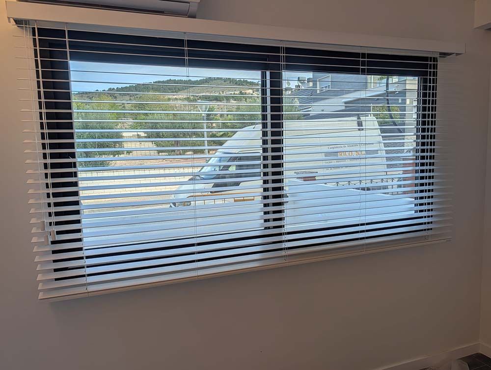 Ventana con persianas blancas parcialmente abiertas, mostrando una vista de una calle y colinas.