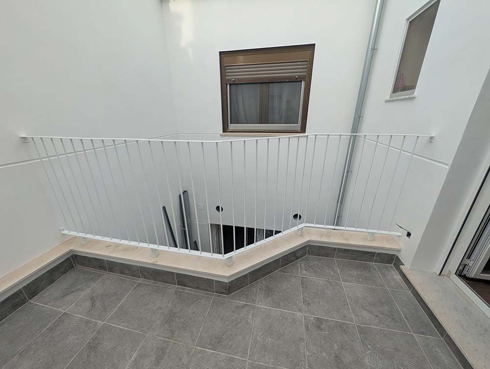 Balcón con barandilla blanca, suelo de baldosas y ventanal con persianas.