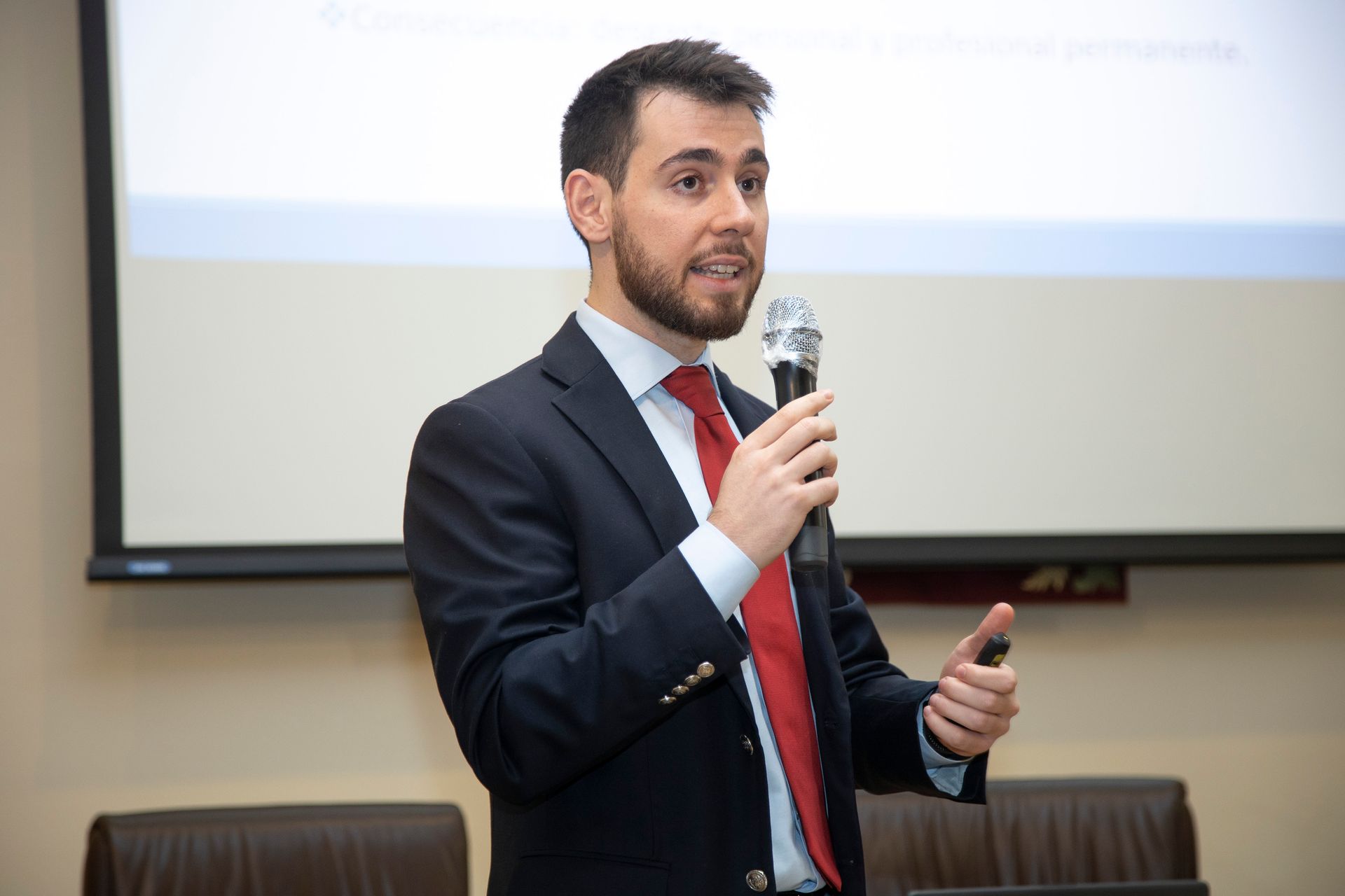 Un hombre con barba y traje habla por un micrófono durante una presentación. Hace un gesto con la mano derecha en una conferencia.
