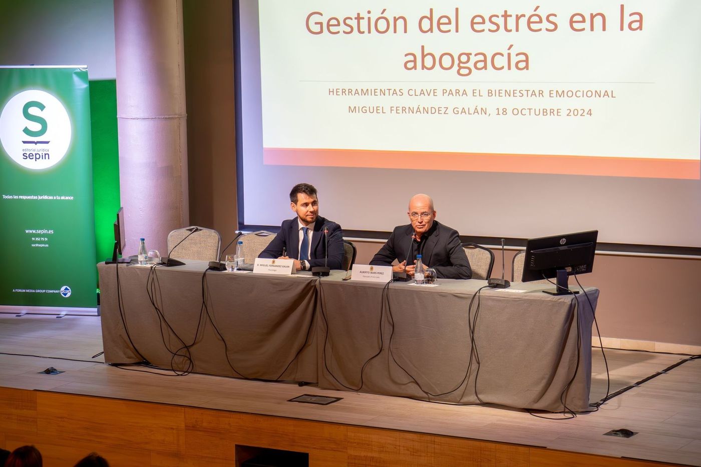 Dos hombres en un escritorio presentan un tema de gestión del estrés para abogados. Una pantalla blanca los respalda.
