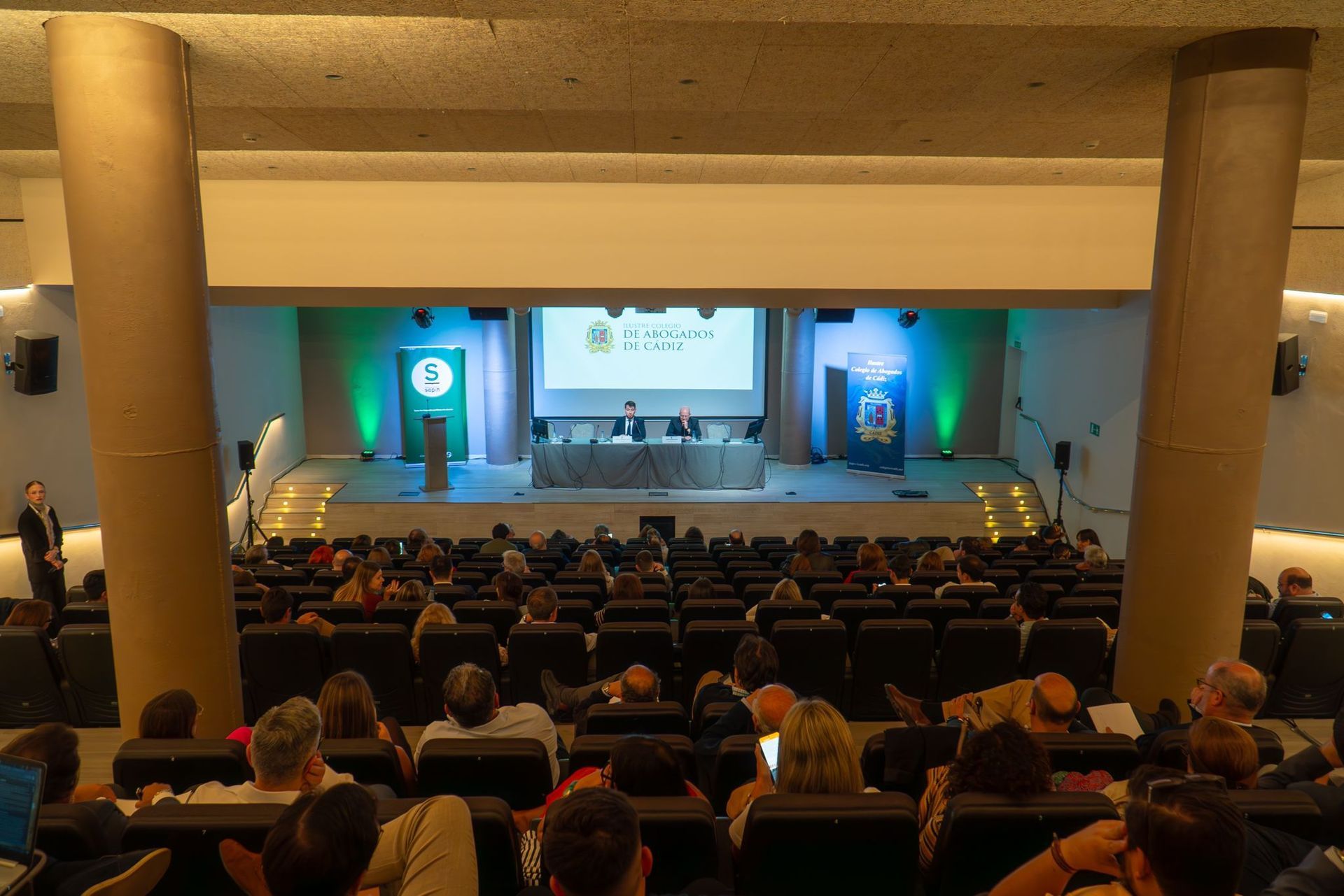 Público sentado en un teatro viendo una presentación en un escenario. El escenario tiene una mesa e iluminación verde.