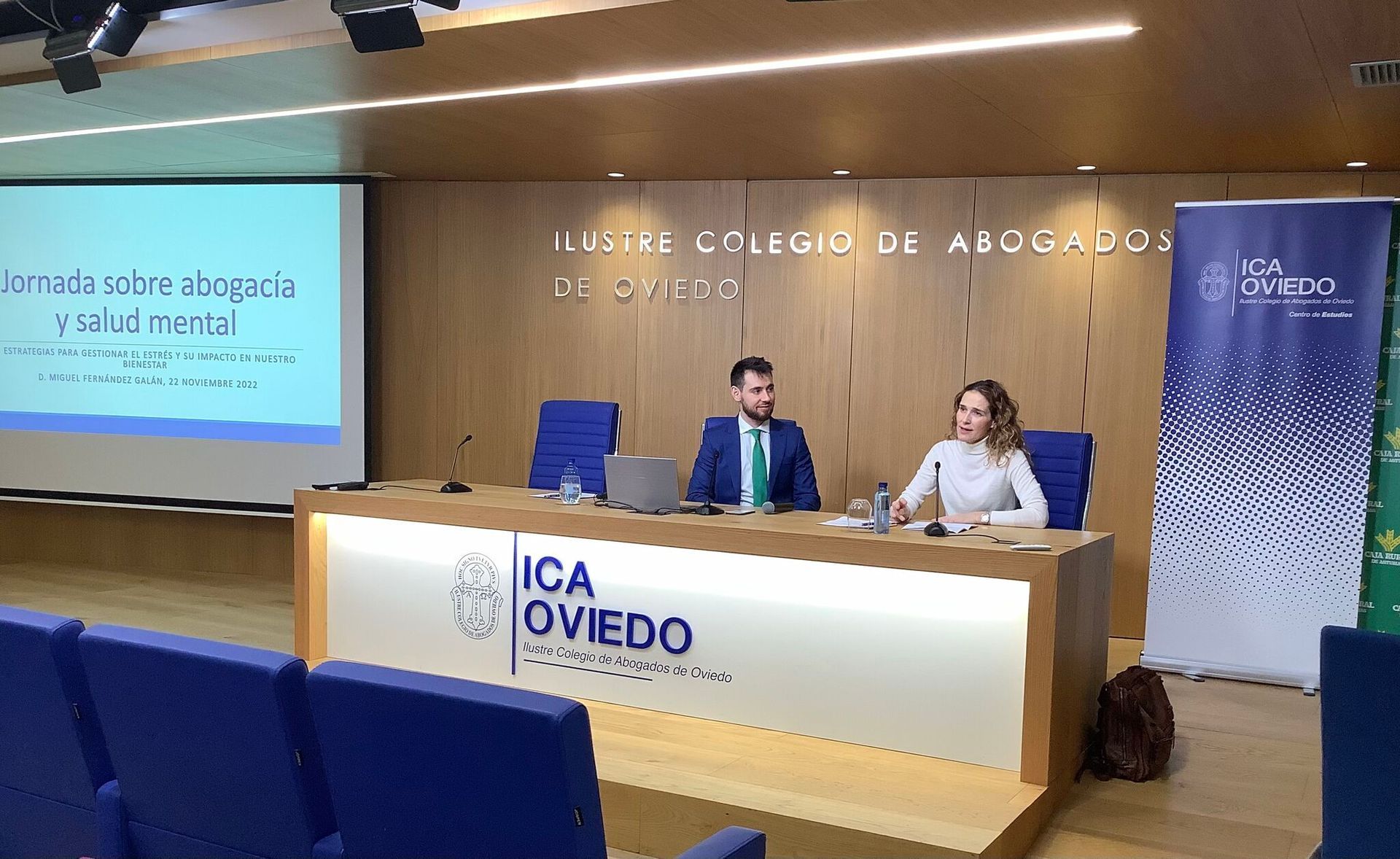 Una presentación en el ICA Oviedo con dos ponentes tras un podio. Una pantalla azul muestra texto sobre defensa y salud mental.