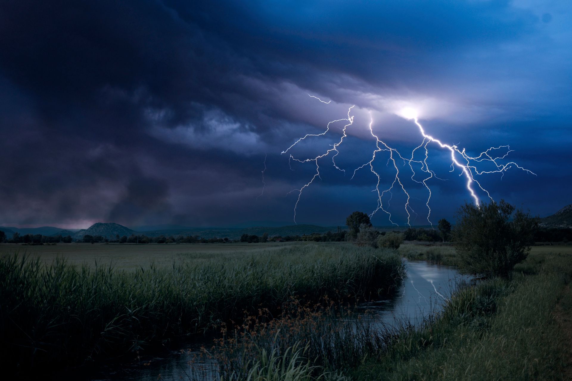 Un rayo cae sobre un campo oscuro y un arroyo, iluminado por un destello blanco brillante. Nubes oscuras llenan el cielo tormentoso.