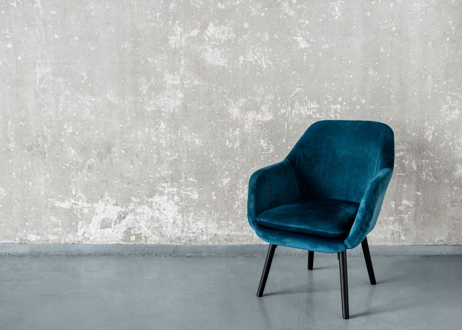 Un sillón de terciopelo verde azulado contra una pared y un suelo de color gris desgastado.