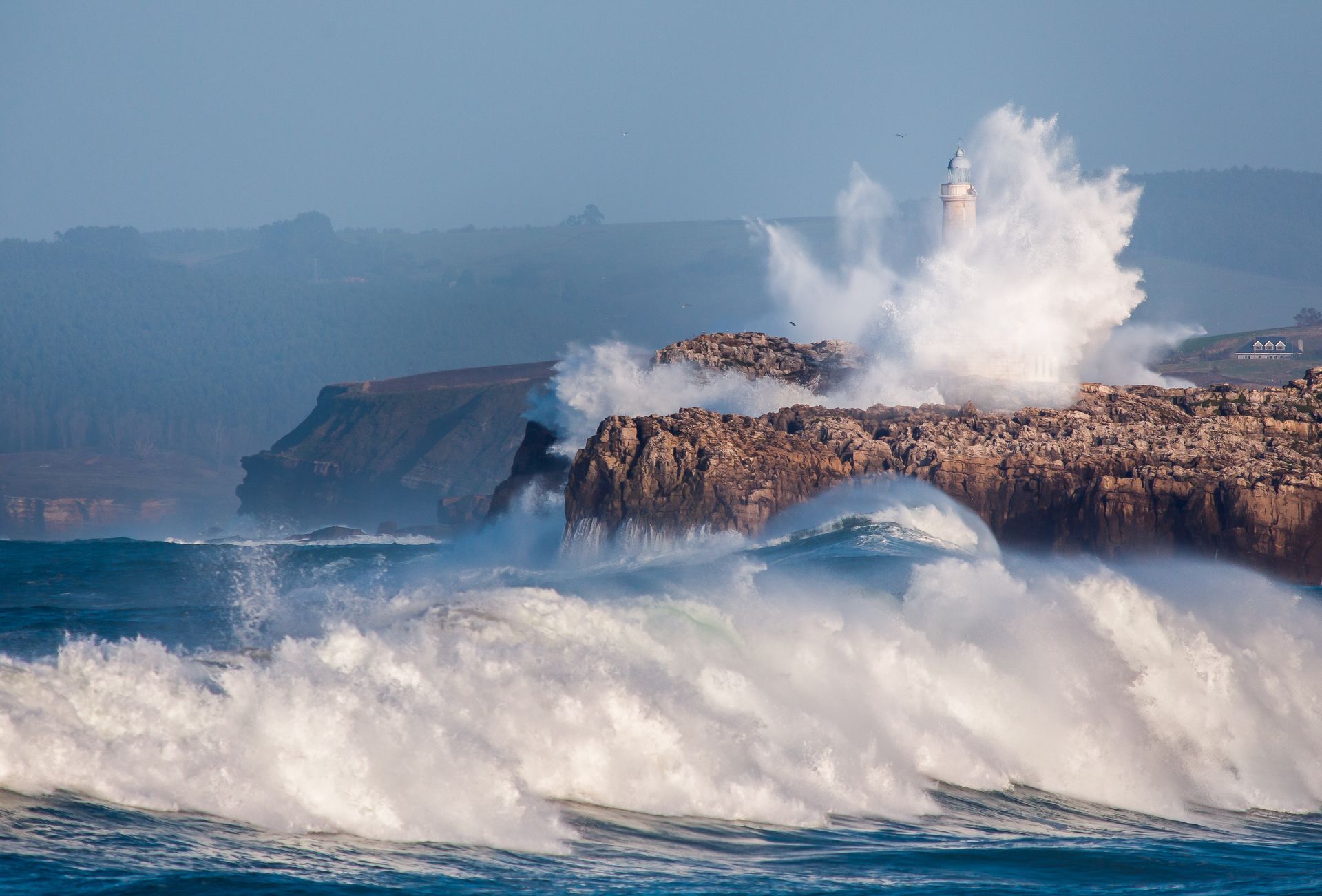 Las olas rompen contra los acantilados rocosos y un faro se yergue mientras el agua explota a su alrededor.