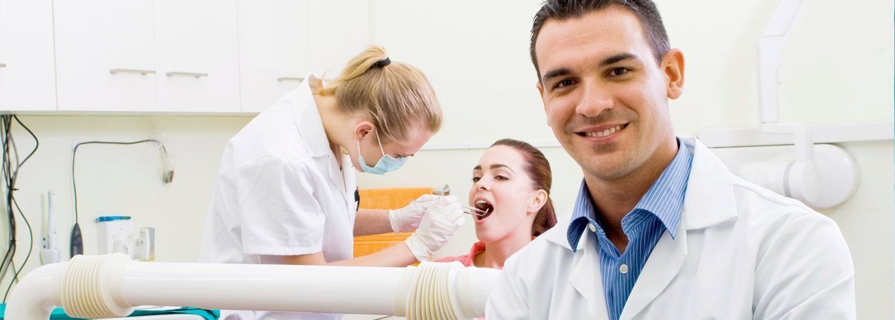 Un dentista sonríe a la cámara mientras un asistente dental trabaja con un paciente en una sala de exámenes.